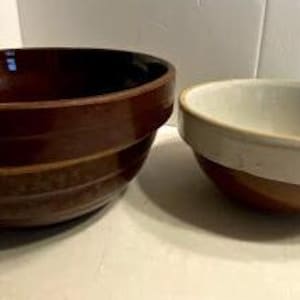 May include: Two vintage brown and white ceramic mixing bowls. The larger bowl has a brown glaze with a white rim and a brown and white striped pattern. The smaller bowl has a white glaze with a brown rim.
