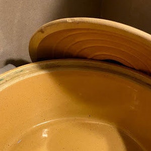 May include: A large, round, terracotta pot with a lid. The pot is a light brown color and has a smooth, glazed finish. The lid is slightly domed and has a small knob on top.