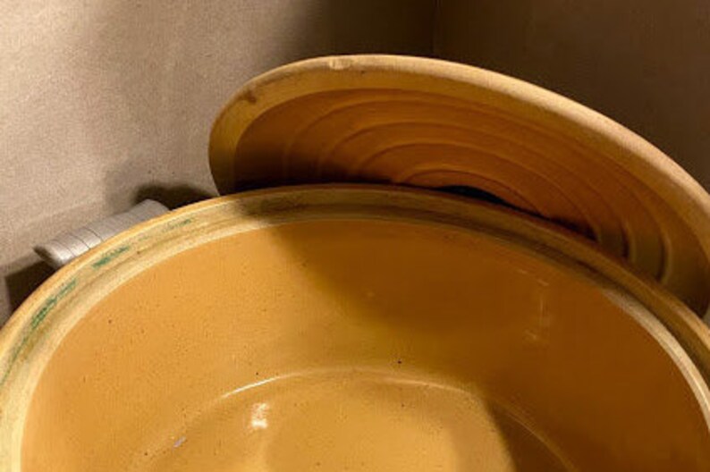 May include: A large, round, terracotta pot with a lid. The pot is a light brown color and has a smooth, glazed finish. The lid is slightly domed and has a small knob on top.