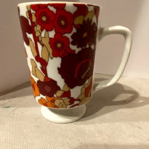 May include: A white ceramic mug with a footed base. The mug features a floral pattern in shades of red, orange, and brown.