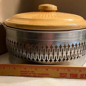 May include: A vintage beige ceramic casserole dish with a metal stand. The stand has a decorative design with a repeating pattern. The dish has a lid.