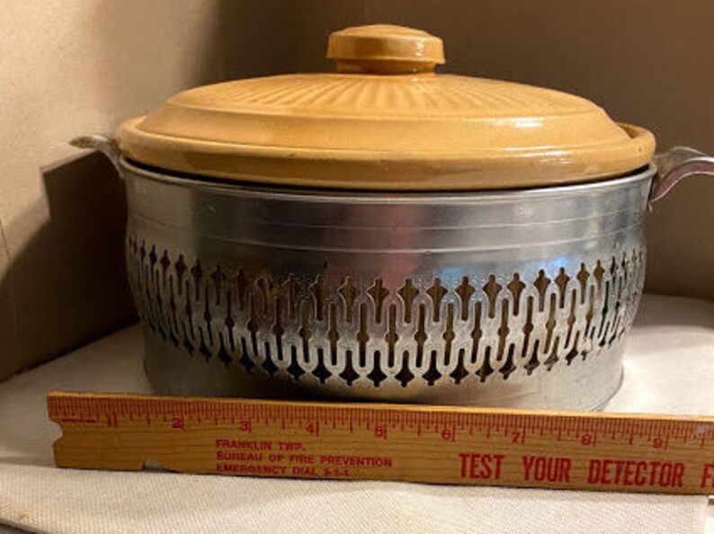 May include: A vintage beige ceramic casserole dish with a metal stand. The stand has a decorative design with a repeating pattern. The dish has a lid.