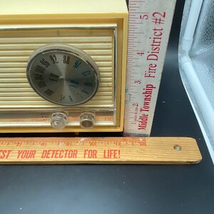 May include: A vintage cream-coloured AM radio with a silver dial and a black knob. The radio has a chrome trim and a black plastic base. The radio is sitting on a wooden ruler with the text "Test Your Detector For Life!" printed on it.