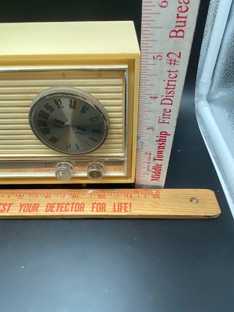 May include: A vintage cream-coloured AM radio with a silver dial and a black knob. The radio has a chrome trim and a black plastic base. The radio is sitting on a wooden ruler with the text "Test Your Detector For Life!" printed on it.