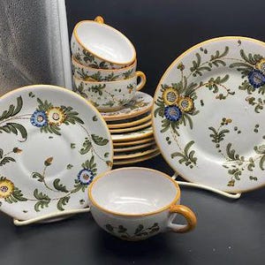 Może przedstawiać: Zestaw vintage włoskich ceramicznych filiżanek do herbaty i spodków z kwiatowymi wzorami w odcieniach niebieskiego, żółtego i zielonego. Filiżanki i spodki mają białe tło z delikatnym wzorem kwiatowym.