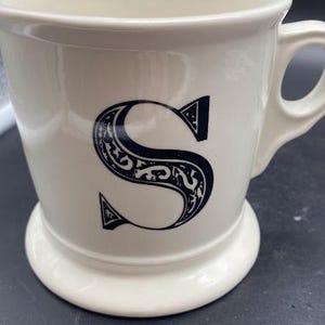 Pu&ograve; includere: Tazza in ceramica bianca con una lettera "S" stilizzata nera sul davanti. La tazza ha un manico e una base larga.
