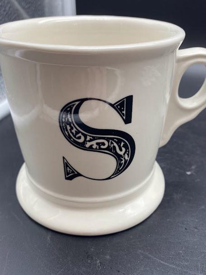 Pu&ograve; includere: Tazza in ceramica bianca con una lettera "S" stilizzata nera sul davanti. La tazza ha un manico e una base larga.