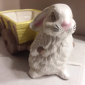 Könnte beinhalten: Eine weiße Keramik-Hasenfigur mit rosa Ohren und schwarzen Schnurrhaaren, die einen gelben und braunen Wagen-Pflanztopf hält.
