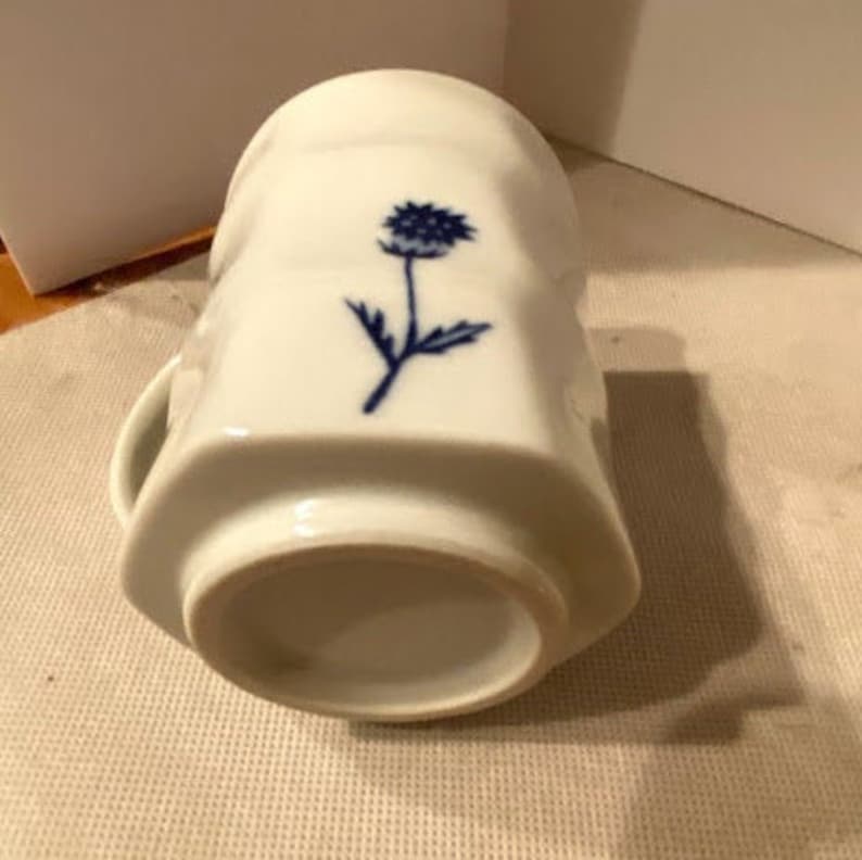 May include: White ceramic mug with a blue floral design. The mug has a rounded bottom and a slightly flared top.