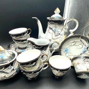 May include: A tea set featuring a teapot, a lidded sugar bowl, a creamer, and several cups with saucers. Each piece is crafted from white ceramic with a black gradient finish, decorated with raised dragon motifs in blue, white, and gold accents.