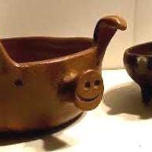 May include: Two handmade ceramic bowls. One bowl is shaped like a pig with a smiling face and the other is a round bowl with three legs.