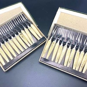 May include: Two boxes of vintage dessert forks. Each box contains a set of twelve forks with silver tines and cream-colored handles. The handles have a gold-colored decorative pattern. The forks are arranged neatly in a black-lined box with a beige lid.
