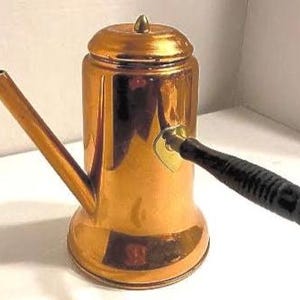Könnte beinhalten: Eine Kaffeekanne aus Kupfer mit einem Holzgriff und einem Messingknopf auf dem Deckel. Die Kanne hat eine glänzende, reflektierende Oberfläche.