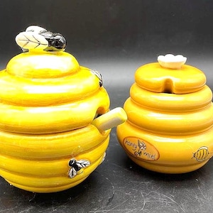 May include: Two yellow ceramic honey pots with lids. One pot is beehive-shaped with a spoon, a bee, and a leaf-topped lid. The other pot is smaller, with a flower-shaped lid and the words "honey miel".