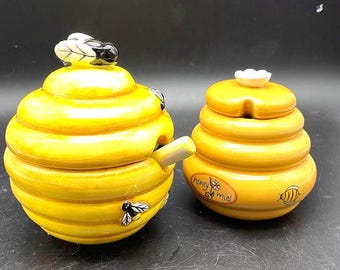 May include: Two yellow ceramic honey pots with lids. One pot is beehive-shaped with a spoon, a bee, and a leaf-topped lid. The other pot is smaller, with a flower-shaped lid and the words "honey miel".