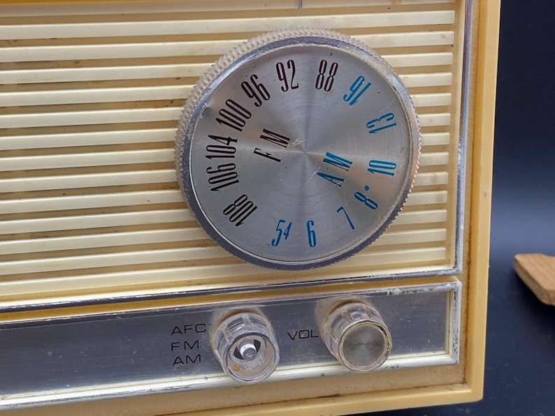 May include: A vintage cream-coloured radio with a silver dial and black lettering. The dial has numbers from 54 to 106 and the words "AM" and "FM". The radio has knobs for "AFC", "FM", "AM", "VOL" and a silver trim around the edge.