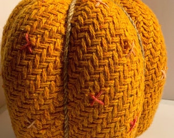 May include: A woven pumpkin decoration with a textured surface and a brown stem. The pumpkin is decorated with small pink and orange stitches.