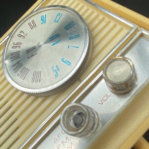 May include: A vintage, cream-coloured AM/FM radio with a silver dial and knobs. The dial has numbers from 54 to 108 and the words "FM" and "AM" are printed on the radio. The knobs are labelled "VOL" and "AFC".