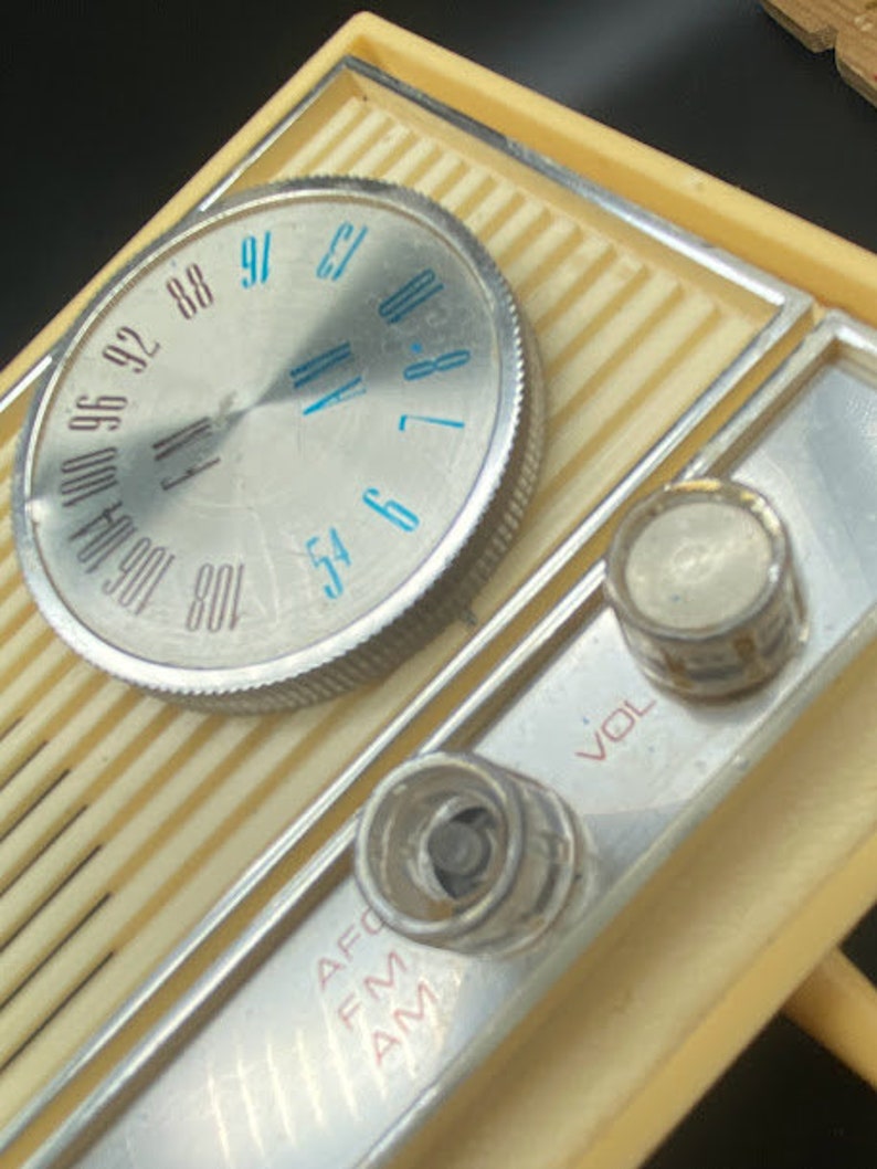 May include: A vintage, cream-coloured AM/FM radio with a silver dial and knobs. The dial has numbers from 54 to 108 and the words "FM" and "AM" are printed on the radio. The knobs are labelled "VOL" and "AFC".