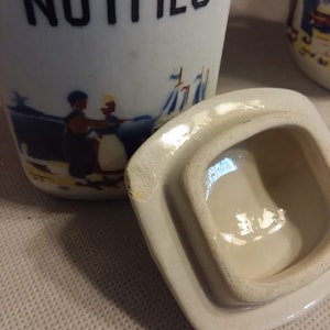 May include: White ceramic spice jar with a blue and yellow scene of people and a sailboat. The lid is off and the jar says "NUTMEG" in black lettering.