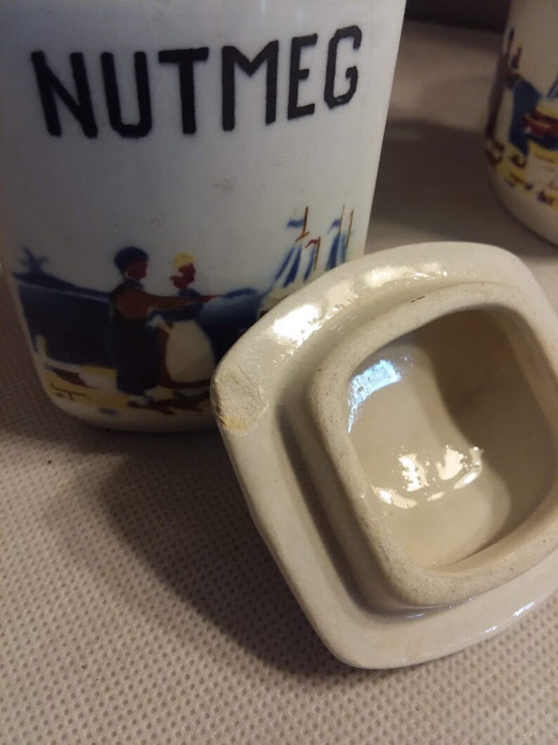 May include: White ceramic spice jar with a blue and yellow scene of people and a sailboat. The lid is off and the jar says "NUTMEG" in black lettering.