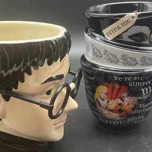 May include: Two ceramic mugs. One mug features a black and white design with the text "Drink Me" and "We're all mad about tea, curious and curiouser, drink me". The other mug features a black and white illustration of Harry Potter with the text "Harry Potter".