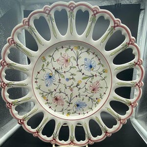 May include: A white ceramic plate with a delicate openwork design and floral pattern in shades of pink, blue, and green. The plate has a scalloped edge and a raised rim.