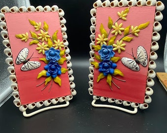 Könnte beinhalten: Zwei kleine rosa gerahmte Bilder mit weißen Muscheln um den Rand. Jedes Bild zeigt ein Blumenmuster mit blauen und gelben Blumen und einem weißen Schmetterling.