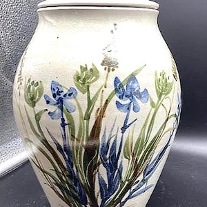 May include: A cream-colored ceramic jar with a lid, adorned with hand-painted blue and green floral designs. The jar has a rounded shape and a small knob on the lid. The floral pattern includes blue iris-like flowers and green foliage.