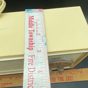 May include: A close-up of a white vintage alarm clock with a black and white dial. The clock is sitting on a wooden surface with a ruler next to it. The ruler is marked with inches and has the text "Middle Township Fire District #2" printed on it.