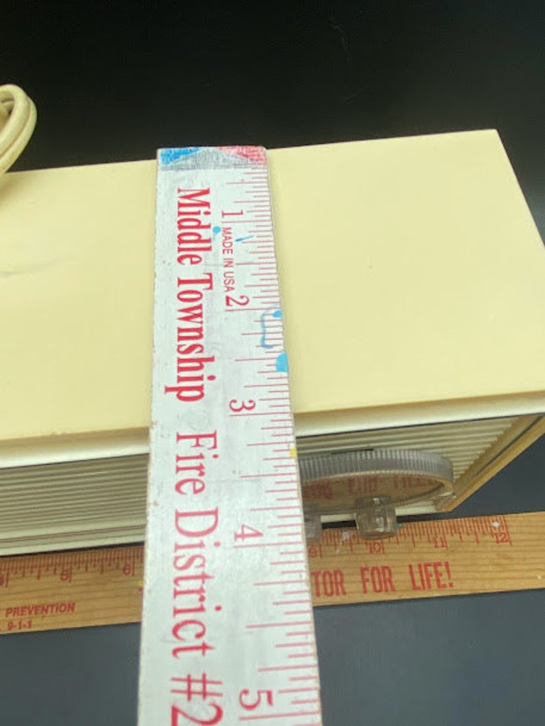 May include: A close-up of a white vintage alarm clock with a black and white dial. The clock is sitting on a wooden surface with a ruler next to it. The ruler is marked with inches and has the text "Middle Township Fire District #2" printed on it.
