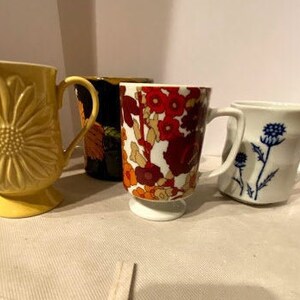May include: Four ceramic mugs with different floral patterns. The mugs are in various colors, including yellow, black, red, and white. The mugs are all in focus and are arranged in a row on a white surface.