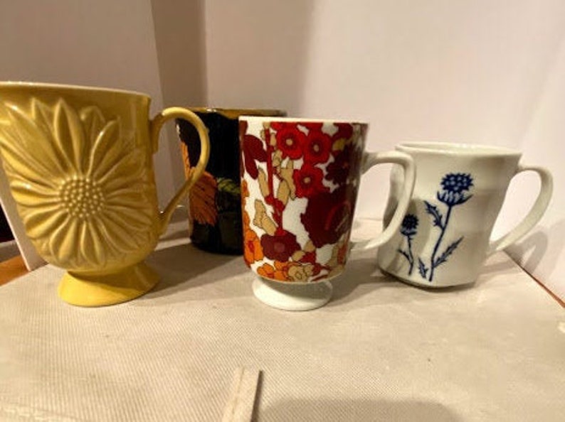 May include: Four ceramic mugs with different floral patterns. The mugs are in various colors, including yellow, black, red, and white. The mugs are all in focus and are arranged in a row on a white surface.