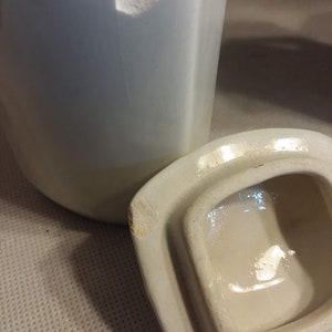 May include: A white ceramic jar with a lid. The jar has a chip on the top edge. The lid is slightly chipped on the edge.