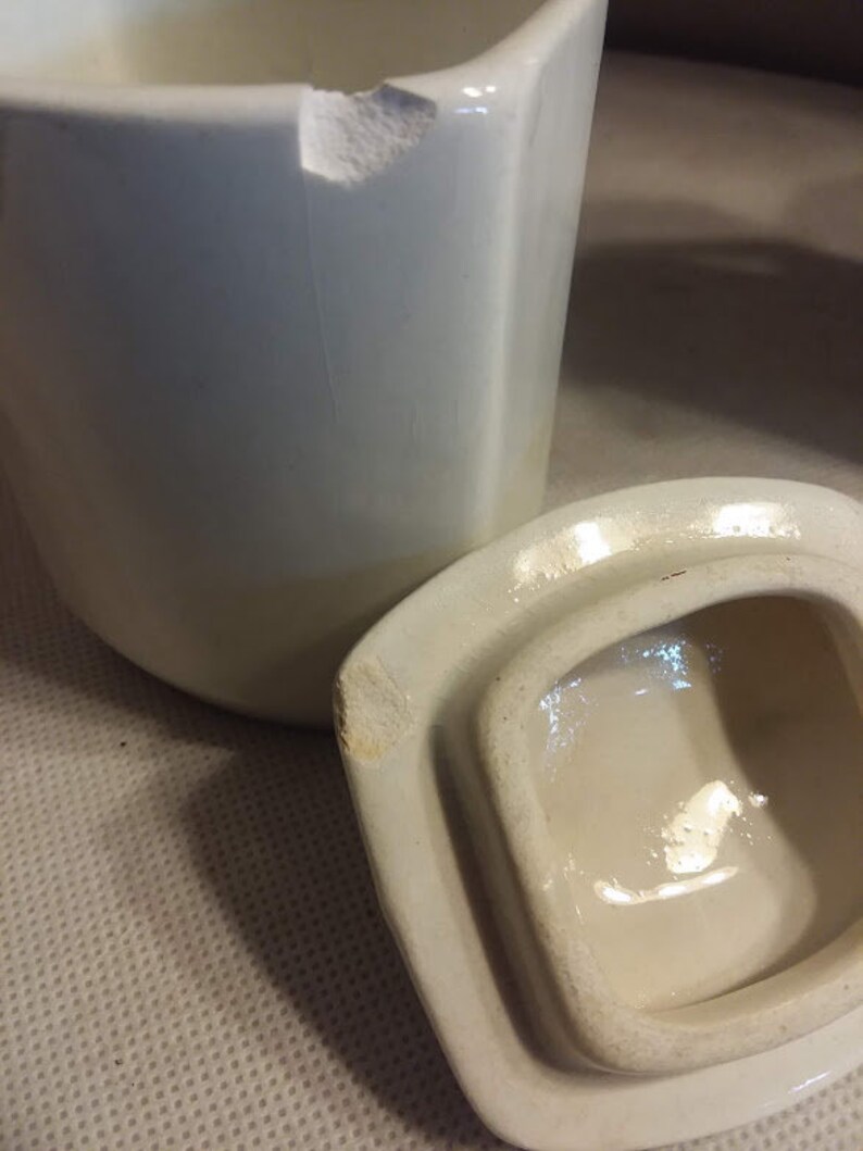 May include: A white ceramic jar with a lid. The jar has a chip on the top edge. The lid is slightly chipped on the edge.