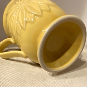 May include: A yellow ceramic mug with a floral design. The mug has a handle and a wide, rounded base.