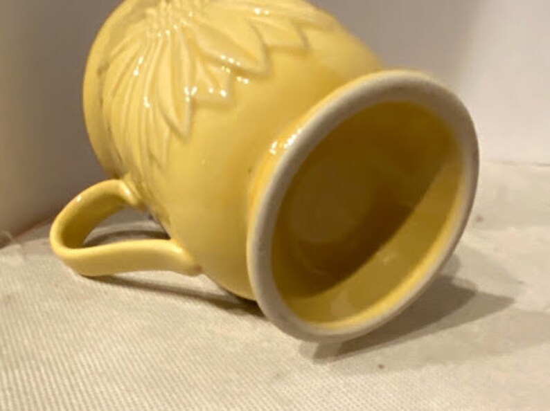 May include: A yellow ceramic mug with a floral design. The mug has a handle and a wide, rounded base.