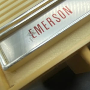 May include: Close-up of a vintage, cream-coloured radio with a silver trim and the word "EMERSON" printed in red letters.