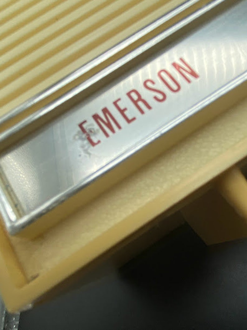 May include: Close-up of a vintage, cream-coloured radio with a silver trim and the word "EMERSON" printed in red letters.