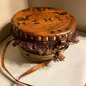May include: A brown leather drum with a fringe border and a woven base. The drum is decorated with a geometric pattern and stars.