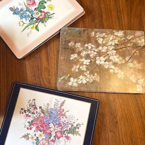 Op de afbeelding: Drie decoratieve placemats met bloemmotieven. Eén heeft een roze rand en een boeket van roze, blauwe en gele bloemen. Een andere heeft witte bloesems op een bruine achtergrond. De derde heeft een marineblauwe rand en een bloemstuk met vogels.