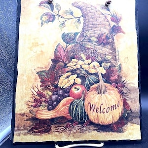 May include: A rectangular slate sign with a fall harvest scene. A cornucopia overflowing with pumpkins, gourds, grapes, and apples is depicted. The word "Welcome" is written on a pumpkin. A brown leather strap is attached for hanging.