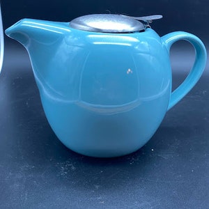 May include: A robin's egg blue ceramic teapot with a stainless steel lid. The teapot has a rounded body, a curved handle, and a spout for pouring. The lid sits on top of the teapot and has a small handle for easy removal. The teapot is designed for brewing tea.