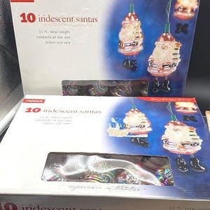 May include: Two white cardboard boxes containing NOMA brand 10 iridescent Santa Claus string lights. Each box features a graphic of the Santa ornaments, text stating 3.35 m total length, and notes that they are for indoor use only.