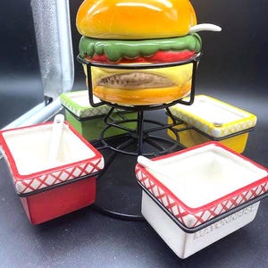May include: A ceramic condiment set featuring a burger-shaped lidded jar and four square bowls with white spoons. The bowls are red, white, green, and yellow, each with a red lattice rim. The white bowl is embossed with the word MAYONNAISE.