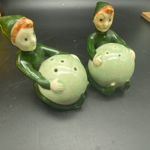 Op de afbeelding: Twee groene en witte keramische elf zout- en peperstrooiers. Elke elf houdt een ronde, groene peperstrooier met gaten in de bovenkant.