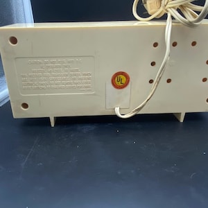 May include: The rear of a white vintage clock radio with a UL safety sticker. The radio has a cord and a series of small holes on the back.