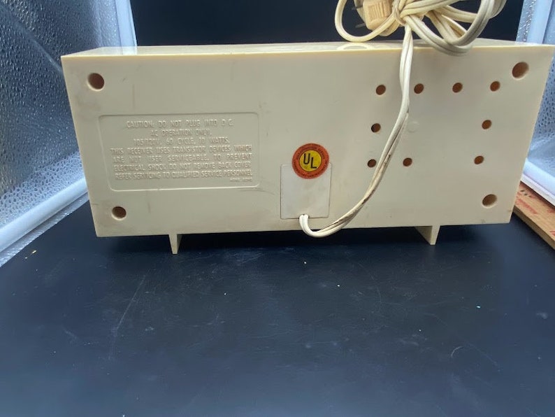 May include: The rear of a white vintage clock radio with a UL safety sticker. The radio has a cord and a series of small holes on the back.
