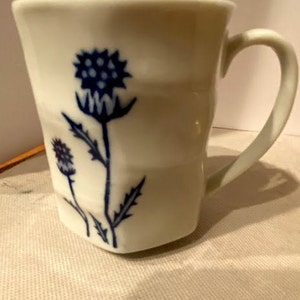 May include: White ceramic mug with a blue and white thistle design. The mug has a rounded handle.