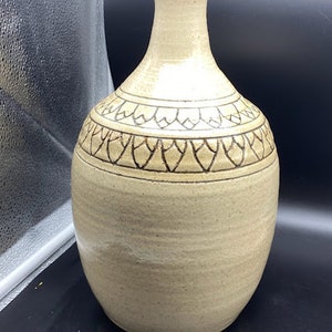 Puede incluir: Un jarrón de cerámica beige con un diseño marrón de hojas estilizadas alrededor del centro. El jarrón tiene un cuerpo redondeado y un cuello estrecho.